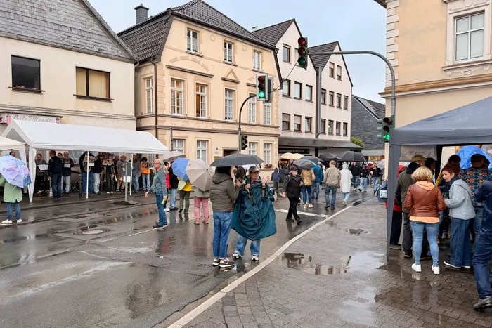 Menschen mit Regenschirmen stehen auf einer nassen Straße vor Fachwerkhäusern und Marktständen. Im Hintergrund ist eine Ampel zu sehen.