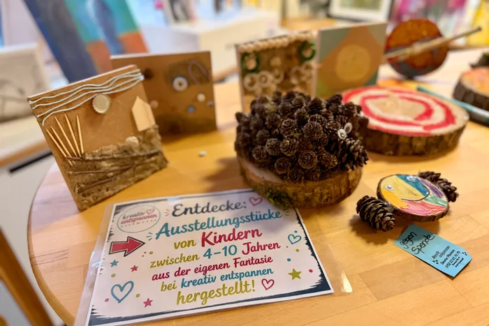 Ausstellungstücke von Kindern zwischen 4 und 10 Jahren, aus eigener Fantasie hergestellt, sind auf einem Holztisch zusammen mit Naturmaterialien ausgestellt. Ein Schild weist auf eine Spendenmöglichkeit hin.