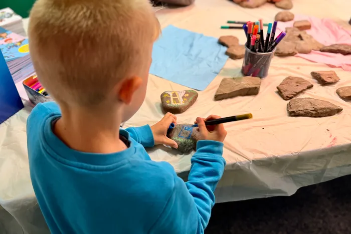 Ein Kind mit blonden Haaren bemalt einen Stein mit einem Pinsel. Auf dem Tisch liegen weitere bemalte Steine und Stifte bereit.