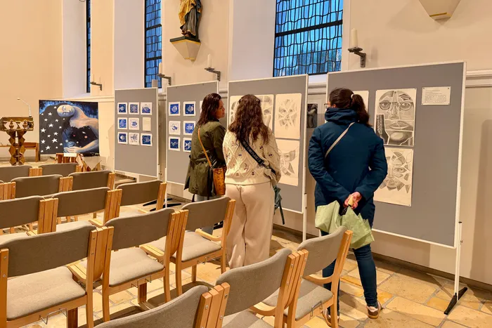 Drei Personen betrachten Zeichnungen und Drucke auf grauen Platten in einer Kirche. Im Hintergrund sind Kirchenbänke und ein hohes Fenster zu sehen.