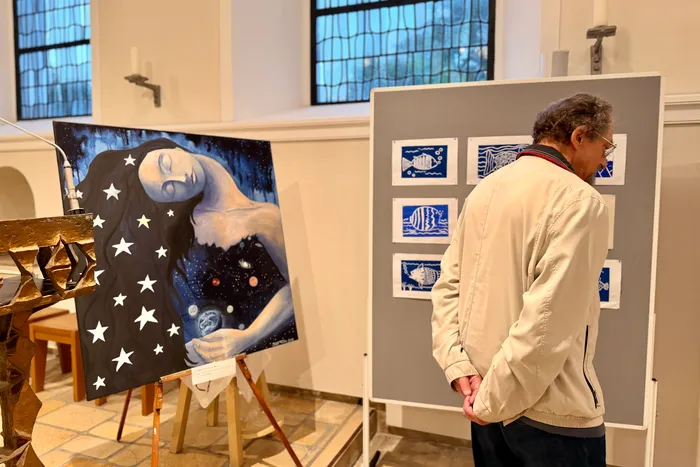 Ein Mann betrachtet in einer hellen Halle Kunstwerke. Links ein Gemälde einer Frau im Weltraum mit Sternen, rechts eine graue Platte mit mehreren kleinen, blauen Drucken von Fischen.