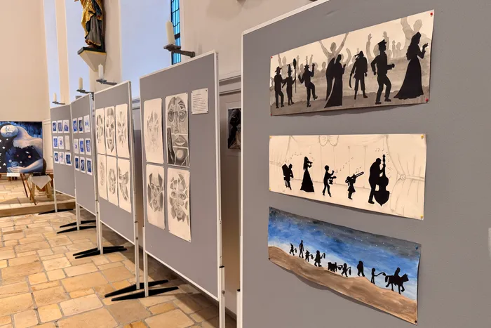 Eine Reihe von grauen Stellwänden zeigt Schwarz-Weiß-Zeichnungen und Silhouettenbilder in einer hellen, kirchenähnlichen Halle mit Fliesenboden. Im Hintergrund ist eine Marienstatue zu sehen.