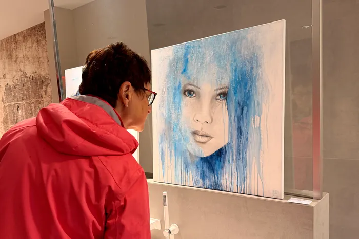 Eine Frau mit roter Jacke betrachtet ein großes, abstraktes Porträt einer Frau mit blauen Augen in einer Ausstellung.