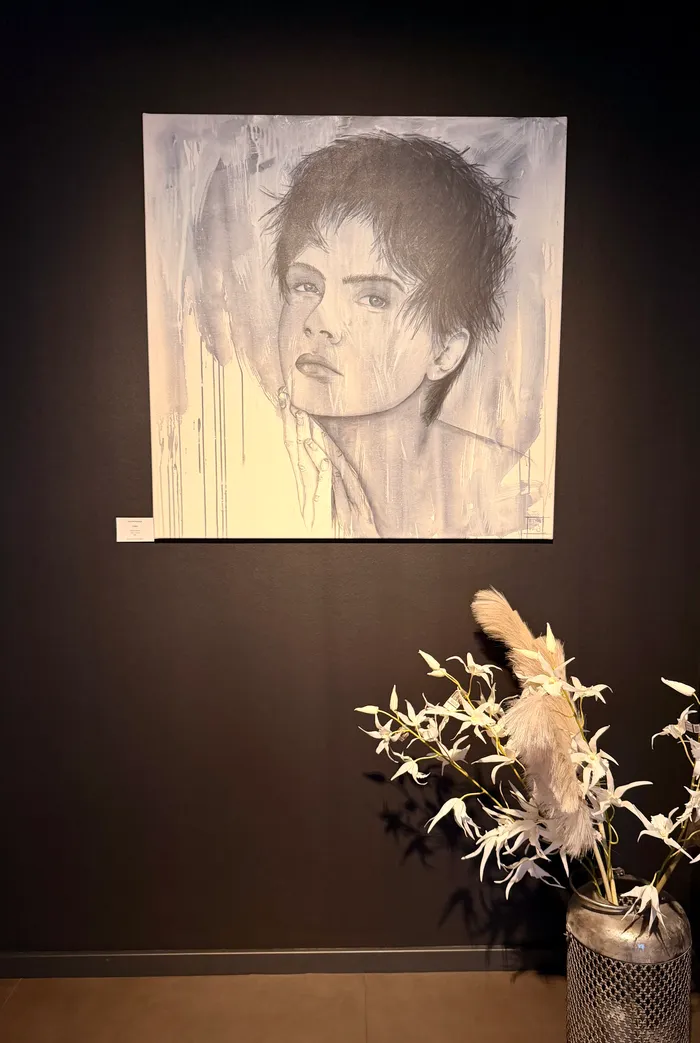 Ein Porträt einer Person mit kurzem, dunklem Haar hängt an einer dunklen Wand. Unter dem Bild steht ein silberfarbener Vasen mit weißen Blumen und einem weichen, flauschigen Zweig.