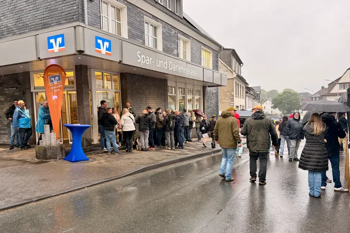 Menschen stehen vor der Sparkasse und warten oder betreten das Gebäude. Im Hintergrund sind weitere Passanten und Gebäude zu sehen.