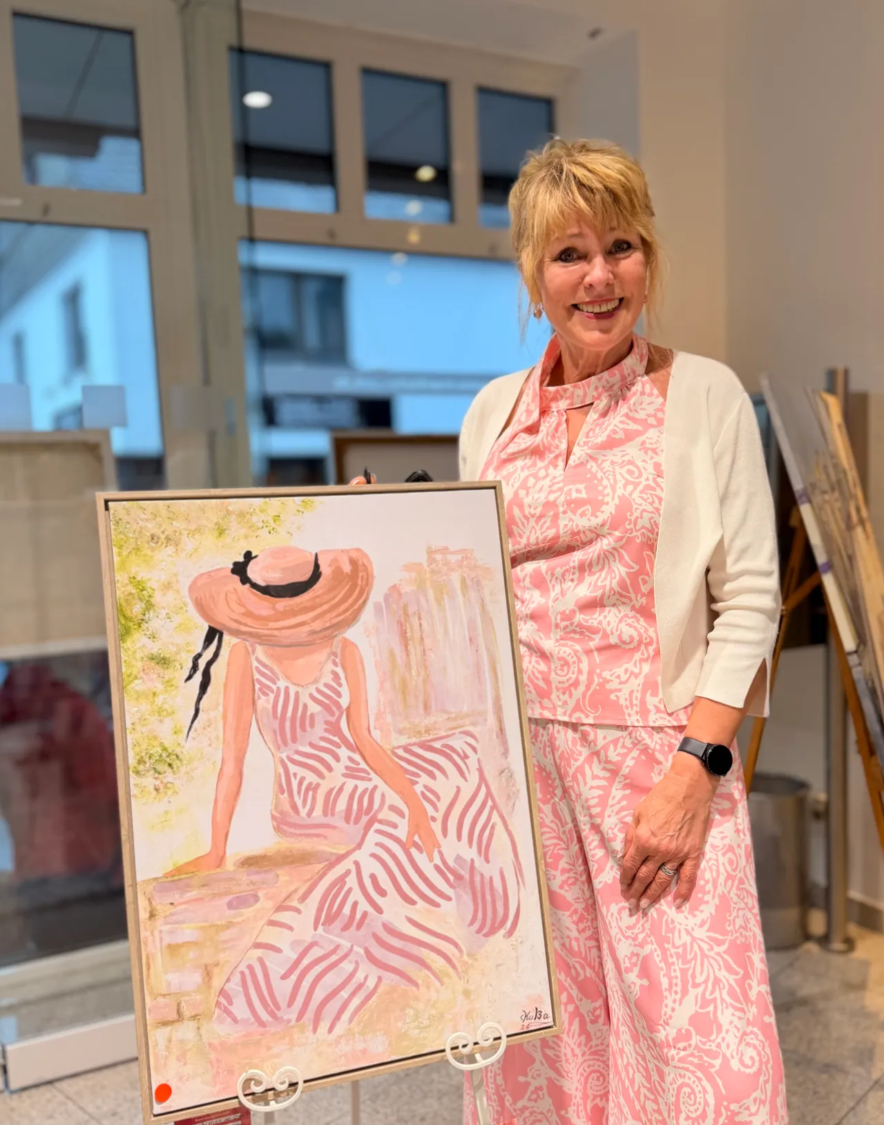 Eine Frau mit blonden Haaren lächelt neben einem Gemälde, das eine Frau mit Hut und gestreiftem Kleid zeigt. Im Hintergrund sind Fenster und weitere Kunstwerke zu sehen.