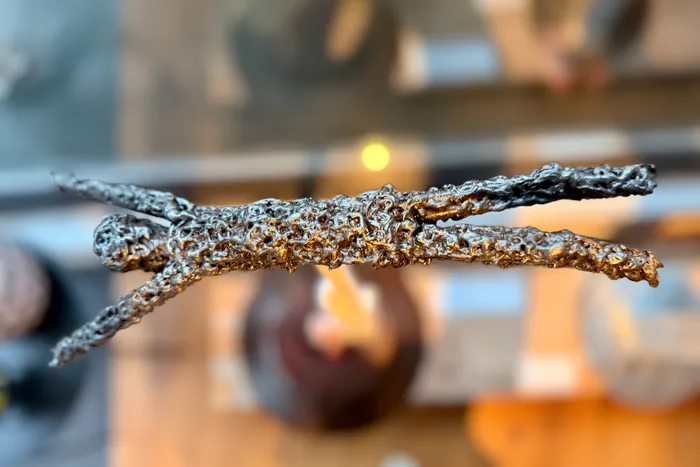 Eine metallische Skulptur in Form einer liegenden menschlichen Figur ist im Fokus. Im Hintergrund sind unscharfe Formen und Lichter zu erkennen.