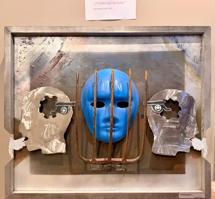 Eine Collage aus einer blauen Maske mit durchgestochenen Metallstreben und zwei metallischen, zerfetzten Maskenfragmenten in einem Bilderrahmen. Die Arbeit trägt den Titel „Friedensgespräche“ und ist mit dem Jahr 2024 signiert.