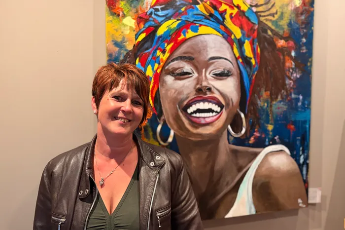 A woman with short, red hair smiles at the camera, standing next to a large, colorful portrait of a Black woman wearing a vibrant headwrap and earrings. The portrait is displayed on a wall in what appears to be a gallery or museum setting.