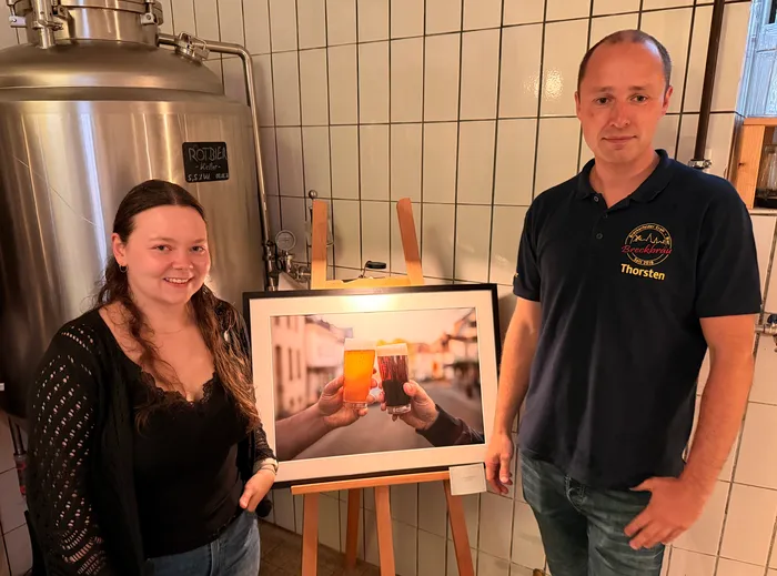 A woman and a man stand side-by-side in a brewery, holding a framed photograph. The photo depicts people raising glasses of beer in a celebratory toast.