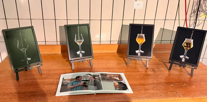 Four framed photographs of different beer glasses are displayed on easels on a wooden table. An open photo book showing images of people enjoying beer is in the foreground.