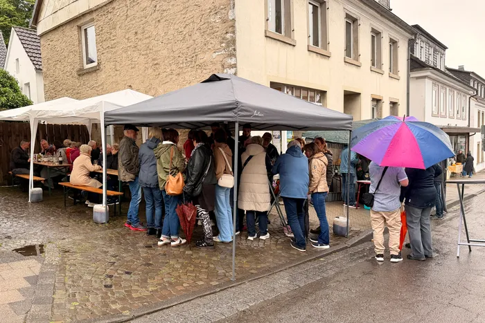 Menschen stehen unter einem Pavillon und warten, während andere an Tischen sitzen und essen. Im Hintergrund sind Gebäude und weitere Personen mit Regenschirmen zu sehen.