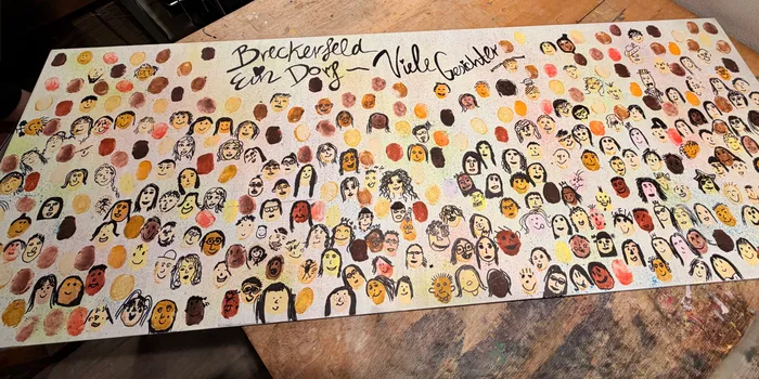Eine lange, rechteckige Leinwand ist mit zahlreichen handgezeichneten Porträts von Menschen unterschiedlichen Alters und mit unterschiedlicher Hautfarbe bedeckt. Darüber steht in geschwungener Schrift 