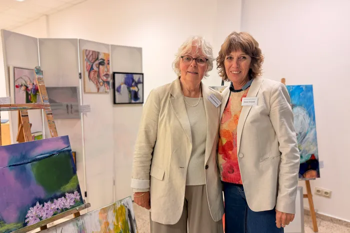 Zwei Frauen mittleren Alters stehen lächelnd vor Gemälden in einer hellen Ausstellungshalle. Beide tragen Blazer und Namensschilder.