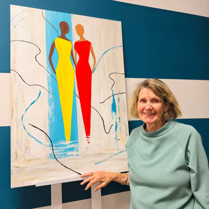 A woman with short blond hair smiles at the camera, standing next to a large, abstract painting. The painting depicts two figures in long dresses, one yellow and one red, against a background of blue and white brushstrokes.