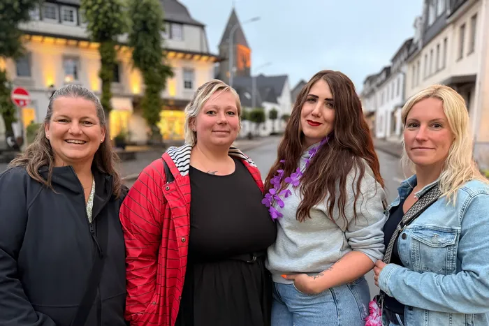 Vier lächelnde Frauen stehen nebeneinander auf einer Straße vor historischen Fachwerkhäusern. Im Hintergrund ist ein Kirchturm zu sehen.