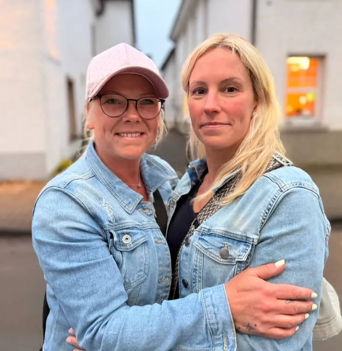 Zwei Frauen umarmen sich vor einer hellen Fassade. Beide tragen Jeansjacken und lächeln in die Kamera.