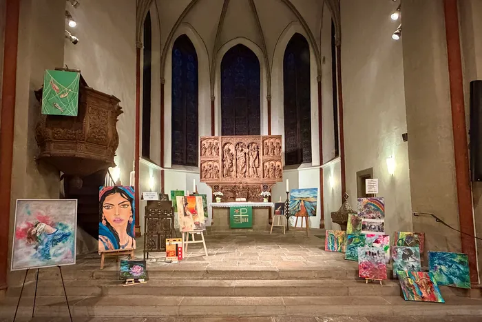 Ausstellungsansicht in einer Kirche mit zahlreichen Gemälden auf Staffeleien und auf dem Boden vor dem Altar. Im Hintergrund sind die hohen Kirchenfenster und die Orgel zu sehen.