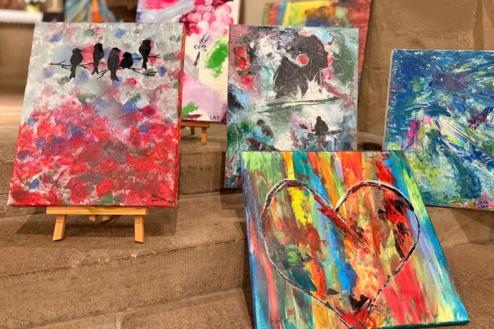 Farbige Gemälde stehen auf Staffeleien und Tischen, darunter eine rote Darstellung von stilisierten Vögeln und ein Herz in leuchtenden Farben. Im Hintergrund sind weitere Kunstwerke zu sehen.