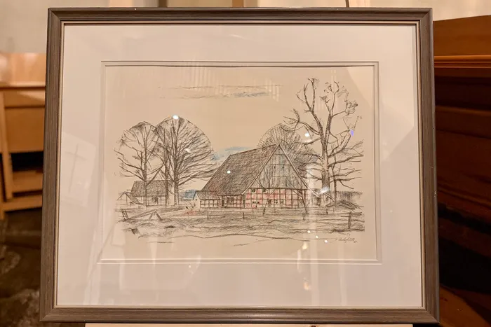 Zeichnung eines Bauernhofs mit rotem Fachwerkhaus und reetgedecktem Dach, umgeben von Bäumen. Im Vordergrund eine Wiese und eine Hecke. Rechts unten eine Signatur.
