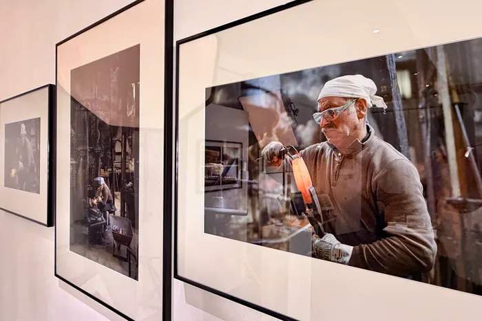 Ein älterer Mann mit Schutzbrille und Kopftuch arbeitet mit einer Flamme an einem Glasobjekt in einer Werkstatt. Das Bild hängt als Teil einer Ausstellung in einem Museum.