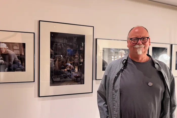 Ein Mann mit Brille und grauem Bart steht vor einer Wand mit gerahmten Schwarz-Weiß-Fotografien in einer Ausstellung. Die Fotos zeigen Handwerker bei der Arbeit.