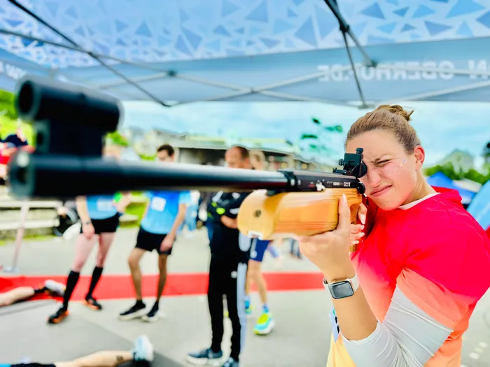 Eine Frau in roter Sportbekleidung zielt mit einem Gewehr auf eine Zielscheibe. Im Hintergrund sind weitere Sportlerinnen und Sportler zu sehen.