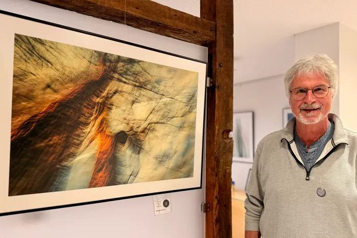 Ein Mann mit grauen Haaren und Brille lächelt neben einem gerahmten, abstrakten Bild eines Pferdekopfes. Im Hintergrund sind weitere Kunstwerke an der Wand zu sehen.