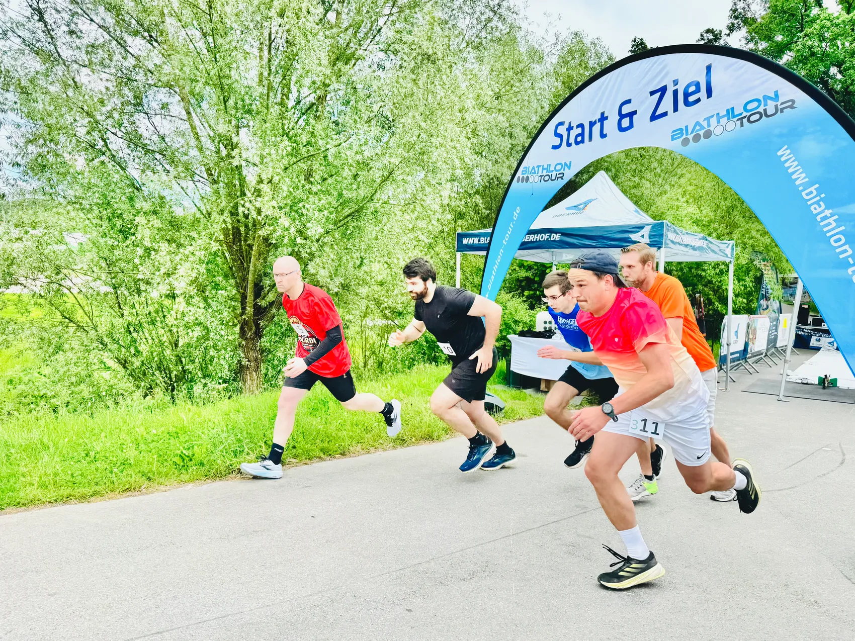 Mehrere Läufer passieren unter einem blauen Bogen mit der Aufschrift „Start & Ziel“ hindurch, während sie an einem Biathlon-Staffel-Challenge-Event teilnehmen. Im Hintergrund sind Bäume und ein Zelt mit Werbeaufschriften zu sehen.