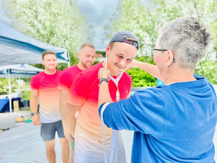 Eine Frau hängt einem lächelnden jungen Mann eine Medaille um, während zwei weitere Männer im Hintergrund stehen. Die Szene spielt im Freien, vermutlich bei einem Sportevent.