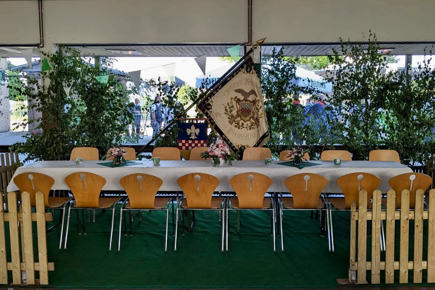 Ein langer Tisch mit weißen Tischdecken und Stühlen steht im Mittelpunkt des Bildes. Im Hintergrund ist eine grüne Wand mit Pflanzen und eine historische Standarte mit Wappen zu sehen.