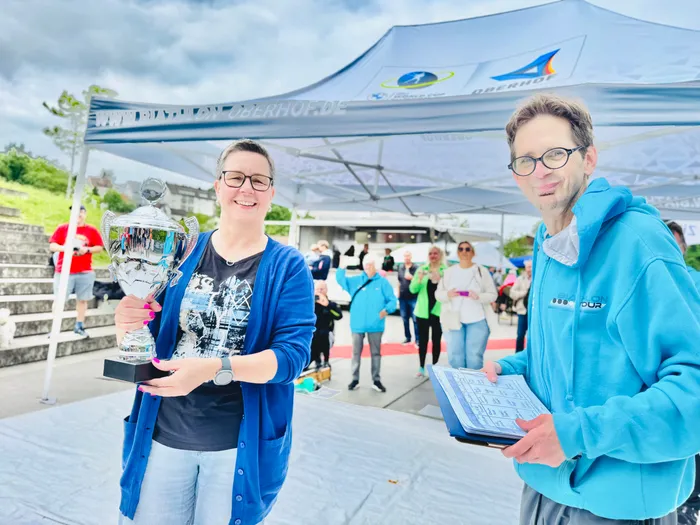 Zwei Personen, eine Frau mit einem Pokal und ein Mann mit Dokumenten, stehen lächelnd vor einem Pavillon mit dem Schriftzug „Biathlon Oberhof“. Im Hintergrund sind weitere Personen und eine Treppe zu sehen.