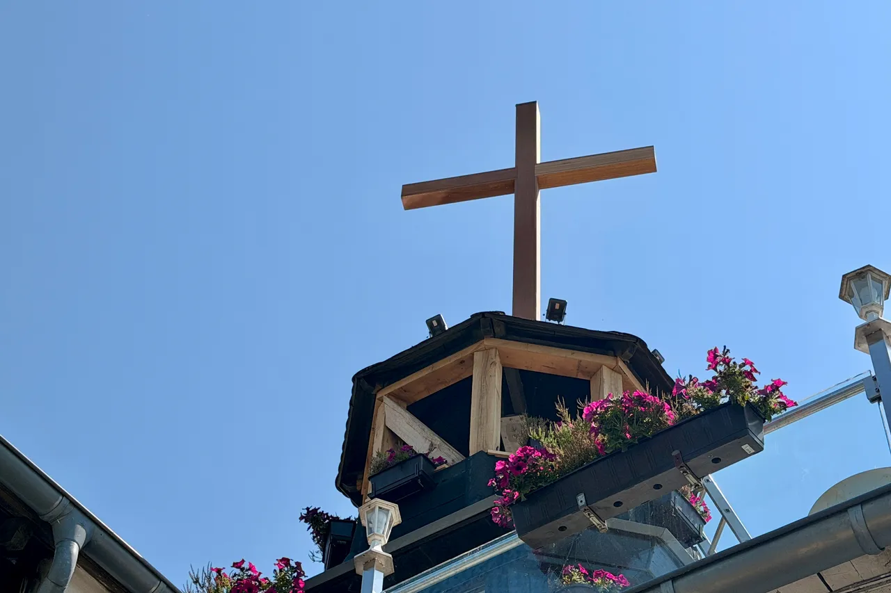 Ein hölzernes Kreuz steht auf dem Dach des „Haus Wengeberg“ in Breckerfeld. Blumenkästen mit pinken Blumen säumen das Dach.