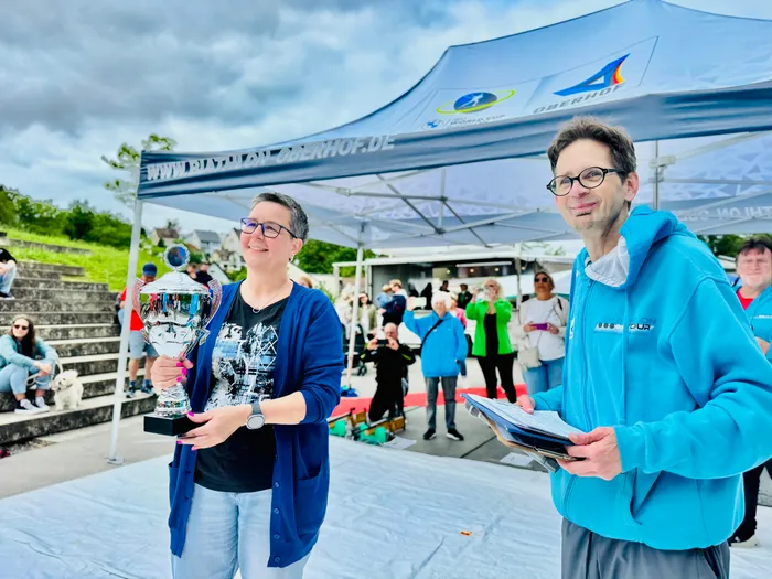 Zwei lächelnde Personen stehen vor einem Zelt mit dem Aufdruck „Biathlon Oberhof“ und halten Pokale bzw. Unterlagen in den Händen. Im Hintergrund sind weitere Sportler und Zuschauer zu sehen.