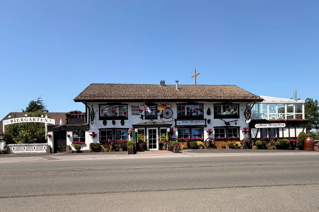Traditional half-timbered building with a steep, thatched roof and a cross on top, displaying signs for 'Haus Wengeberg' and a 'Biergarten'. Decorated balconies and old wagon wheels add to the rustic charm of the building.
