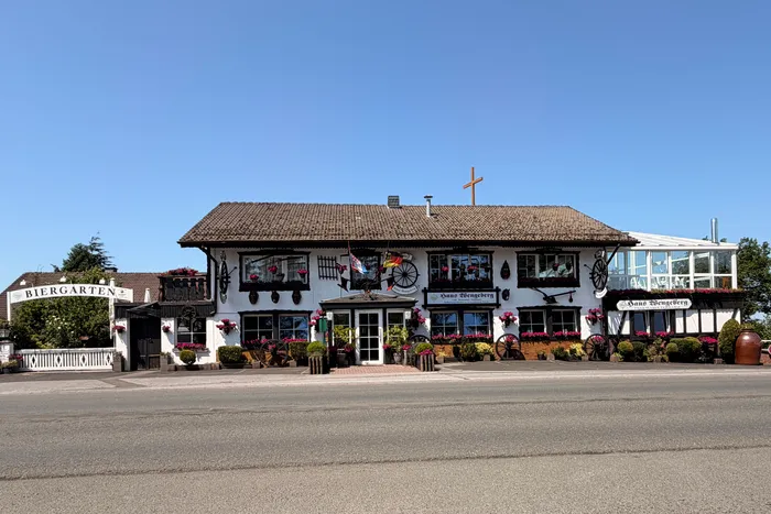 Traditional half-timbered building with a steep, thatched roof and a cross on top, displaying signs for 'Haus Wengeberg' and a 'Biergarten'. Decorated balconies and old wagon wheels add to the rustic charm of the building.