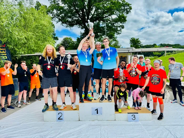 Sieger der 4. Biathlonstaffel-Challenge stehen auf einem Podest mit den Stufen 1, 2 und 3. Der Mann auf der ersten Stufe hält eine Trophäe hoch, während die anderen Athleten um ihn herum Medaillen und Pokale tragen. Im Hintergrund ist eine Landschaft mit Bäumen und einem bewölkten Himmel zu sehen.