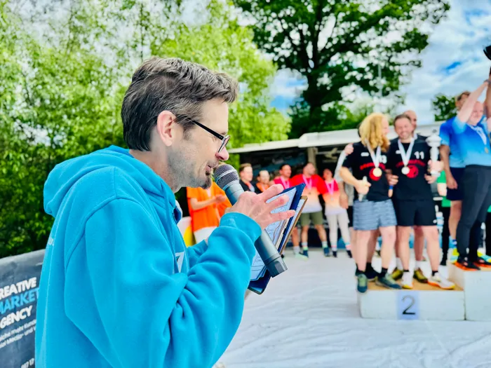 Ein Mann mit Brille spricht in ein Mikrofon, während er Notizen von einem Blatt Papier liest. Im Hintergrund steht ein Podest mit der Nummer 2 und weitere Personen.