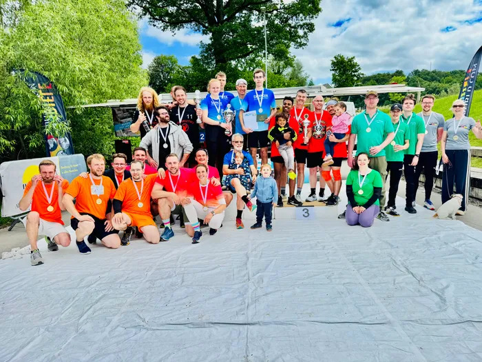 Eine Gruppe von etwa 20 Personen steht auf einer provisorischen Bühne mit Teppichboden vor einer grünen Kulisse. Viele tragen Medaillen um den Hals und halten Pokale oder Urkunden. Im Hintergrund sind Bäume und ein Gebäude zu sehen.