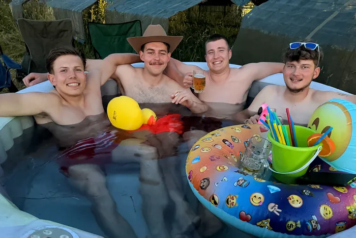 Vier junge Männer sitzen lächelnd in einem aufblasbaren Pool. Einer trägt einen Cowboyhut und hält ein Glas in der Hand, während ein anderer eine gelbe Quietscheente hält. Getränke und Snacks stehen auf einem bunten Schwimmring im Wasser.