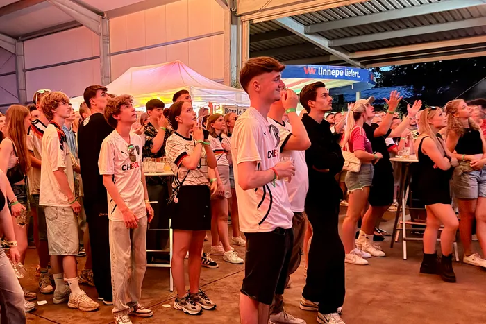 Junge Menschen stehen auf einem Festgelände und schauen aufgeregt nach oben. Einige tragen Trikots der deutschen Fußballnationalmannschaft und Deutschlandflaggen. Im Hintergrund sind Zelte und Werbebanner zu sehen.