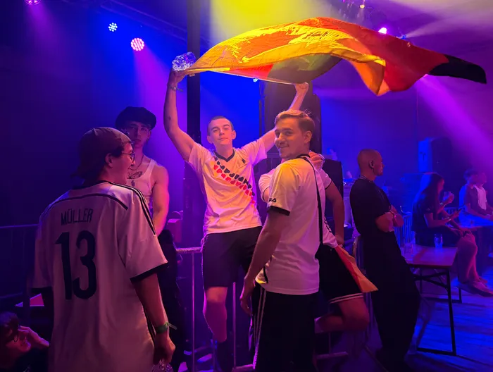 Junge Menschen feiern auf einem Scheunenfest. Ein Mann hält eine große Deutschlandflagge hoch, während andere in Fußballtrikots stehen und lächeln. Im Hintergrund ist eine Bühne mit farbiger Beleuchtung zu sehen.