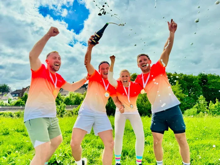 Drei Männer und eine Frau mit Medaillen und Sportkleidung stehen auf einer Wiese und versprühen Champagner. Sie jubeln und heben die Arme in die Luft.
