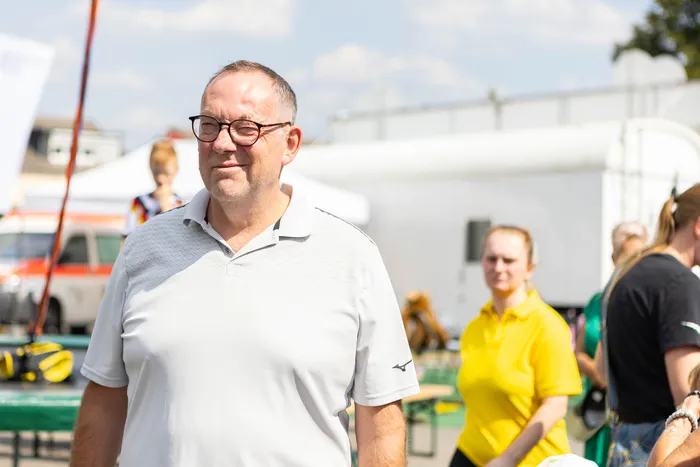 Ein Mann mit Brille und weißem Poloshirt steht im Freien vor Menschen und weißen Zelten.