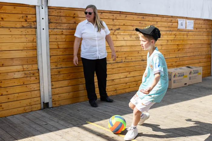 Ein Junge in hellblauem T-Shirt und schwarzer Kappe spielt mit einem bunten Fußball auf einer Holzterrasse.