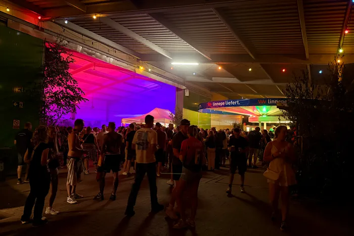 Menschen stehen und gehen auf einem belebten Festivalgelände mit bunten Lichtern und Getränke- und Imbissständen im Hintergrund.