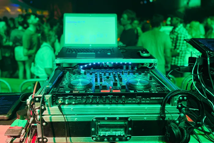 DJ-Pult mit Mischpult, Laptop und Kopfhörern vor einer Menschenmenge in einem grün beleuchteten Raum.