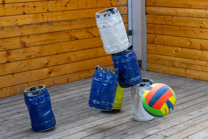 Ein bunter Volleyball in der Luft vor mehreren blauen und weißen Metallfässern auf einem Holzdeck.