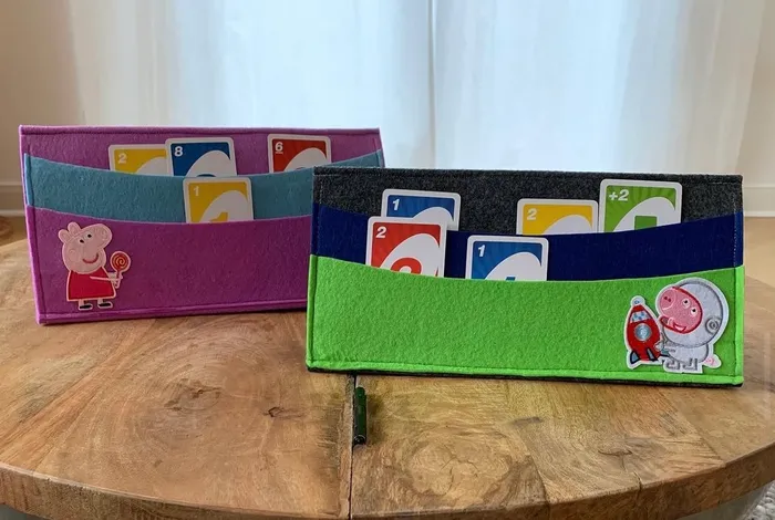 Zwei Spielkartenhalter aus Stoff in Pink und Grün mit Motiven aus der Kinderserie Peppa Pig stehen auf einem Holztisch. Sie dienen dazu, Spielkarten aufrecht zu halten.