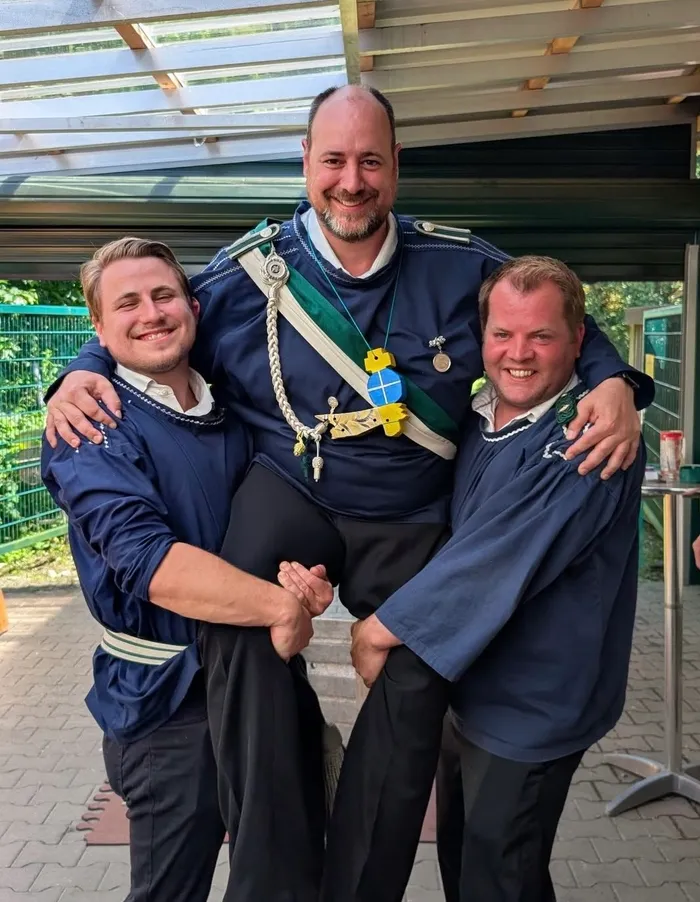 Drei Männer posieren eng umschlungen für ein Foto. Der Mann in der Mitte trägt eine dunkle Uniform mit vielen Abzeichen und Medaillen, während die beiden anderen Männer ähnliche, aber einfachere blaue Jacken tragen.